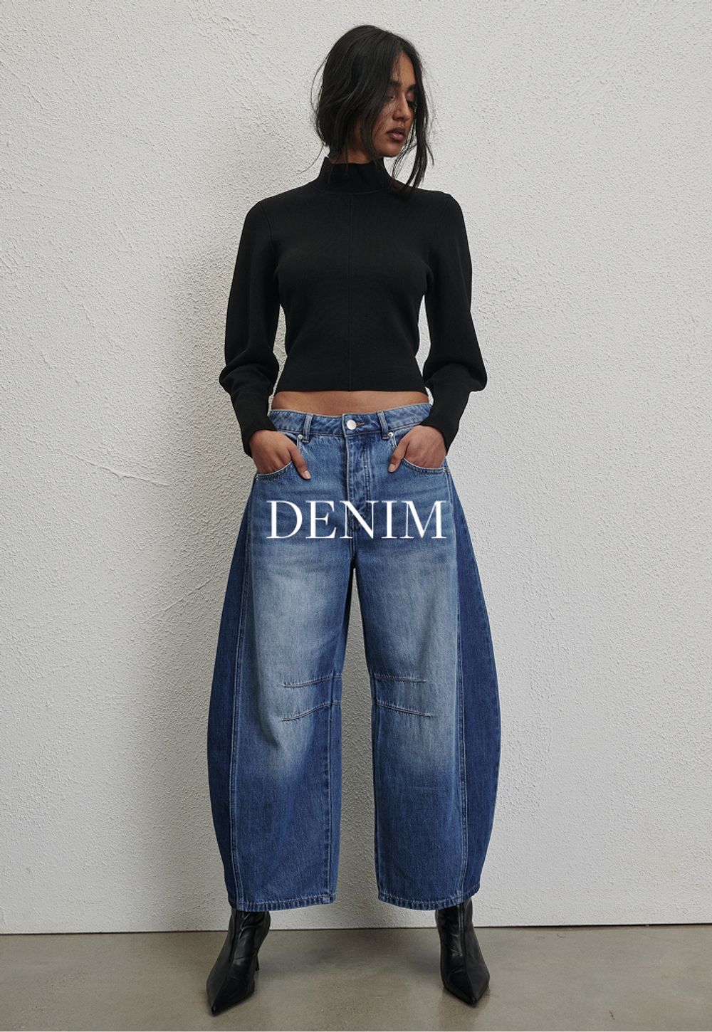 Woman posing in front of a white wall wearing a jeans and a black long sleeve top with text that states "denim"