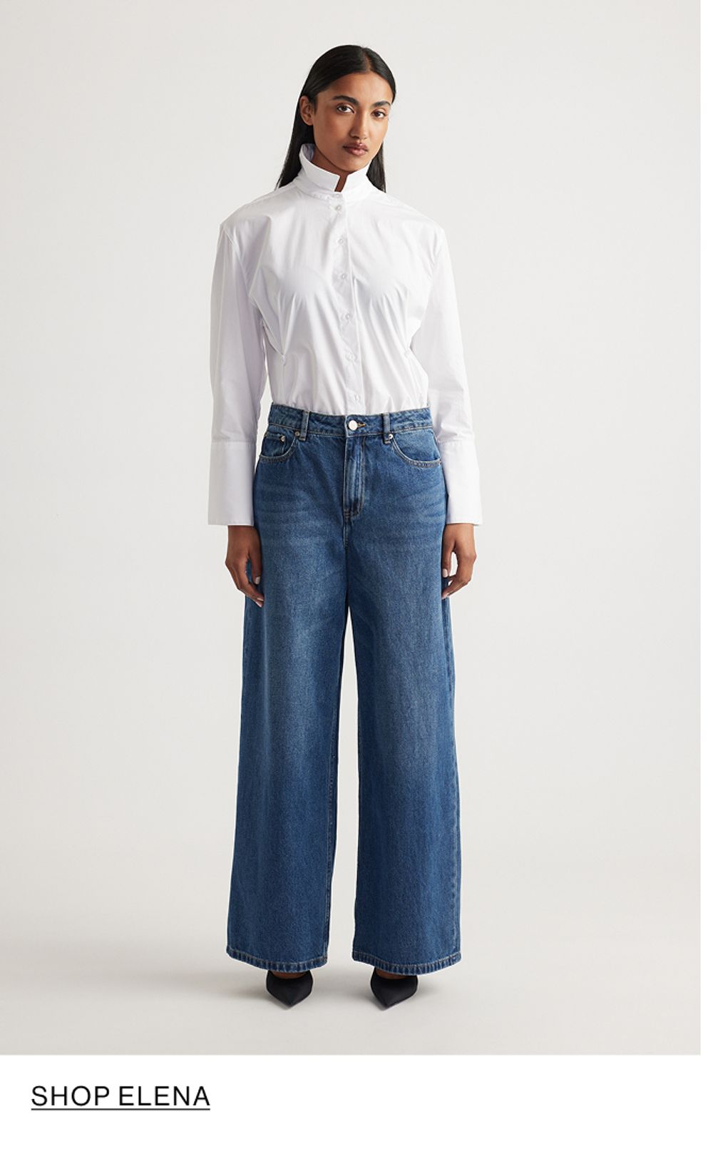 Woman standing in front of a white wall wearing a white shirt and wide jeans, with overlaid text that reads “Shop Elena.”