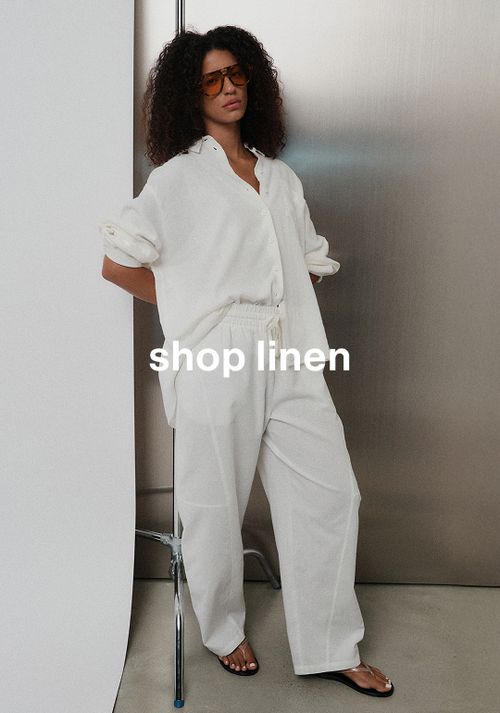 Model in matching white linen shirt and long pants, standing casually against a metal wall with one hand behind her back. Text reads "Shop linen"
