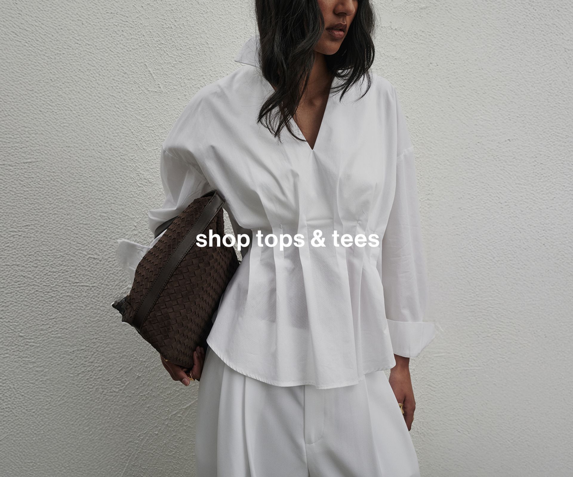Model in a crisp white pleated shirt with a collar, holding a sleek black bag, standing against a minimalist white wall and gazing downward.