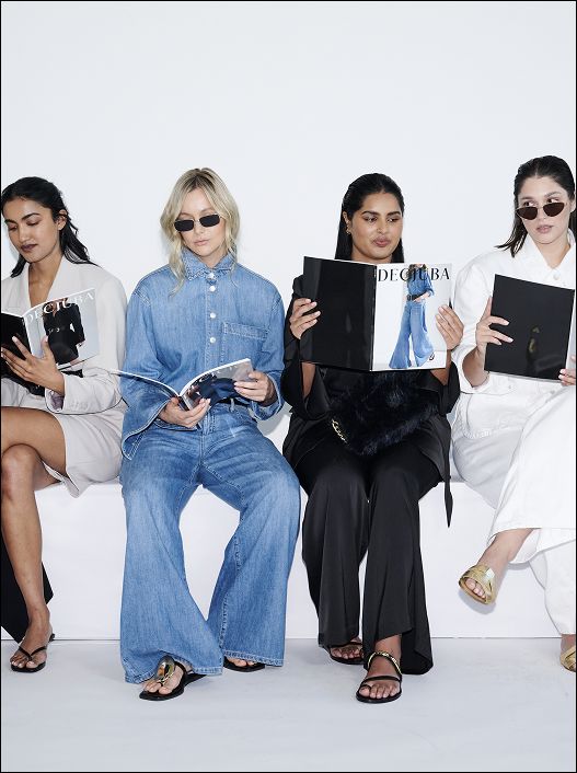 Four women sitting in a row, each wearing fashionable outfits and sunglasses, reading fashion magazines. The mood is stylish and contemporary.