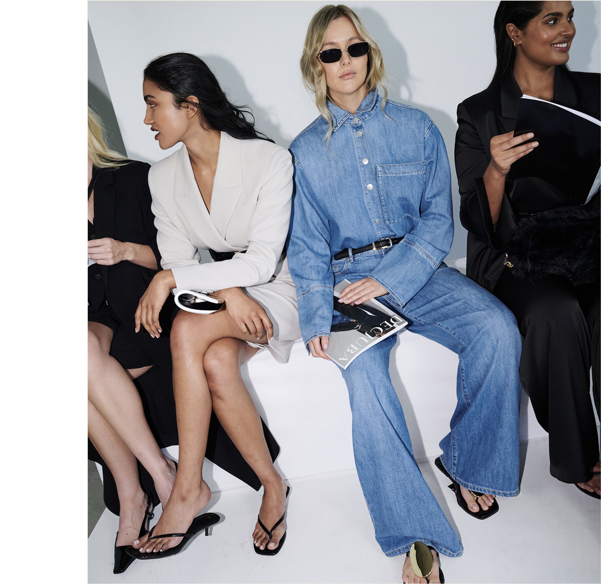 A group of women sitting on a bench, wearing stylish clothing. One woman in denim with sunglasses holds a magazine.