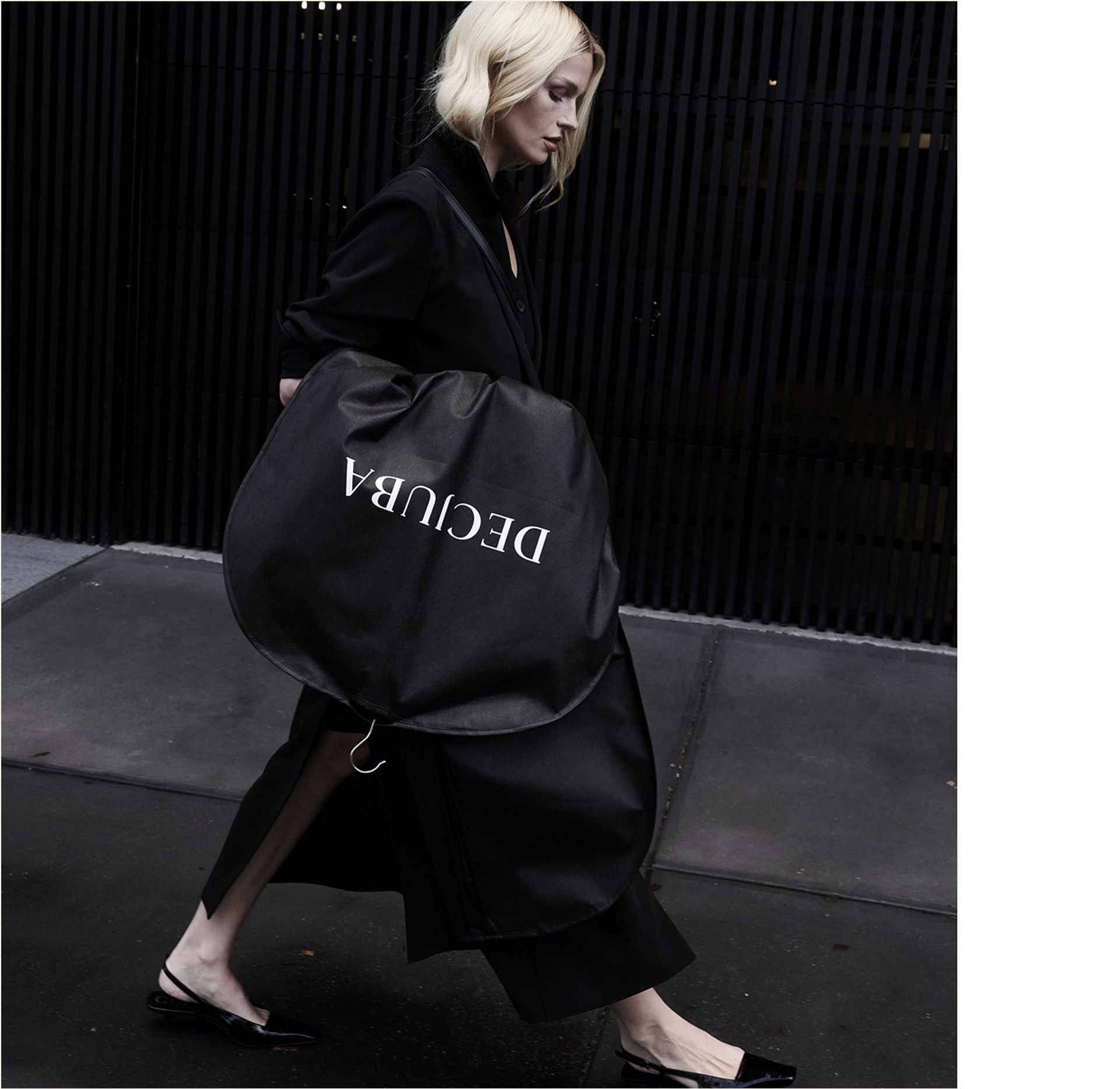 A woman in a sleek black outfit walks outside with a large, round black coat bag labelled "DECUBA"