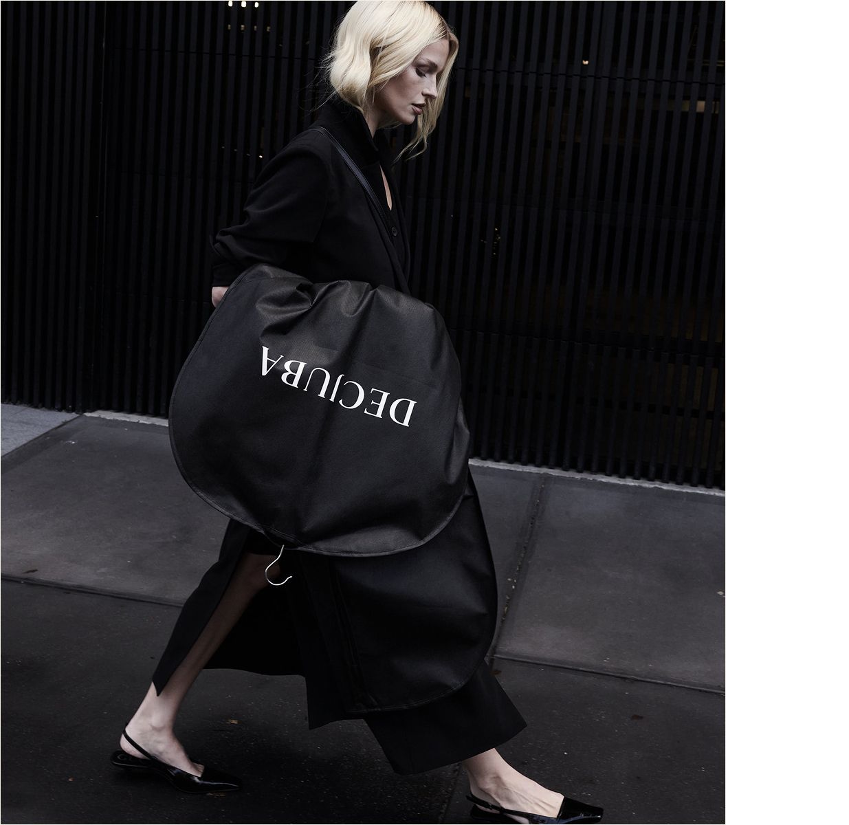 A woman in a sleek black outfit walks outside with a large, round black coat bag labelled "DECUBA"