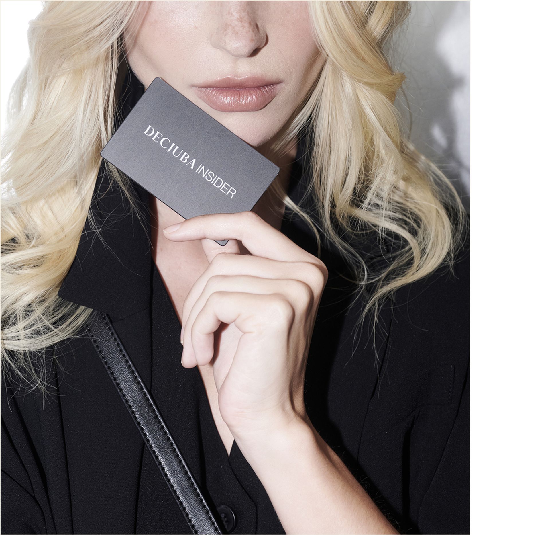 A woman with wavy blonde hair holds a dark "DECJUBA INSIDER" loyalty card, wearing a black outfit.