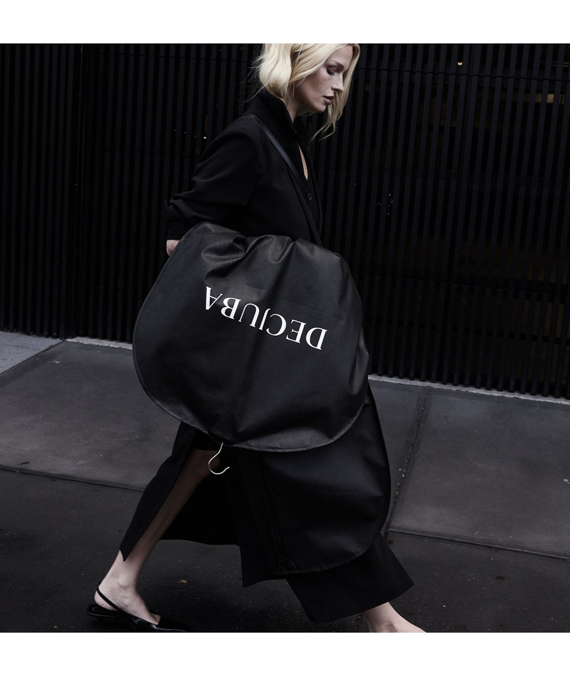 A woman in a sleek black outfit walks outside with a large, round black coat bag labelled "DECUBA"