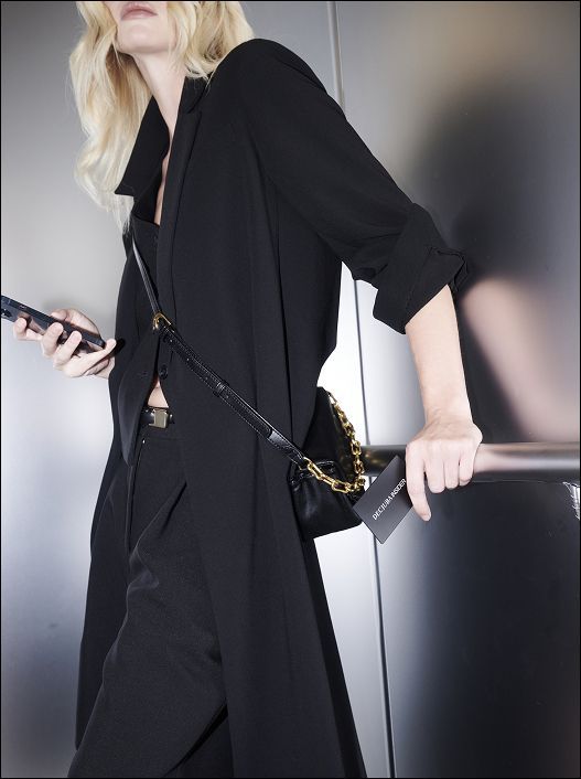 A person with blonde hair in a black outfit and coat leans against an elevator wall, holding a smartphone and a small black bag with a gold chain.