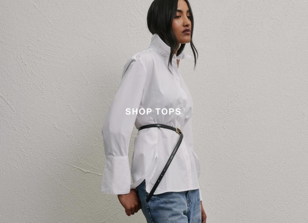 Model in a white blouse and jeans standing against a neutral wall – Shop Tops.