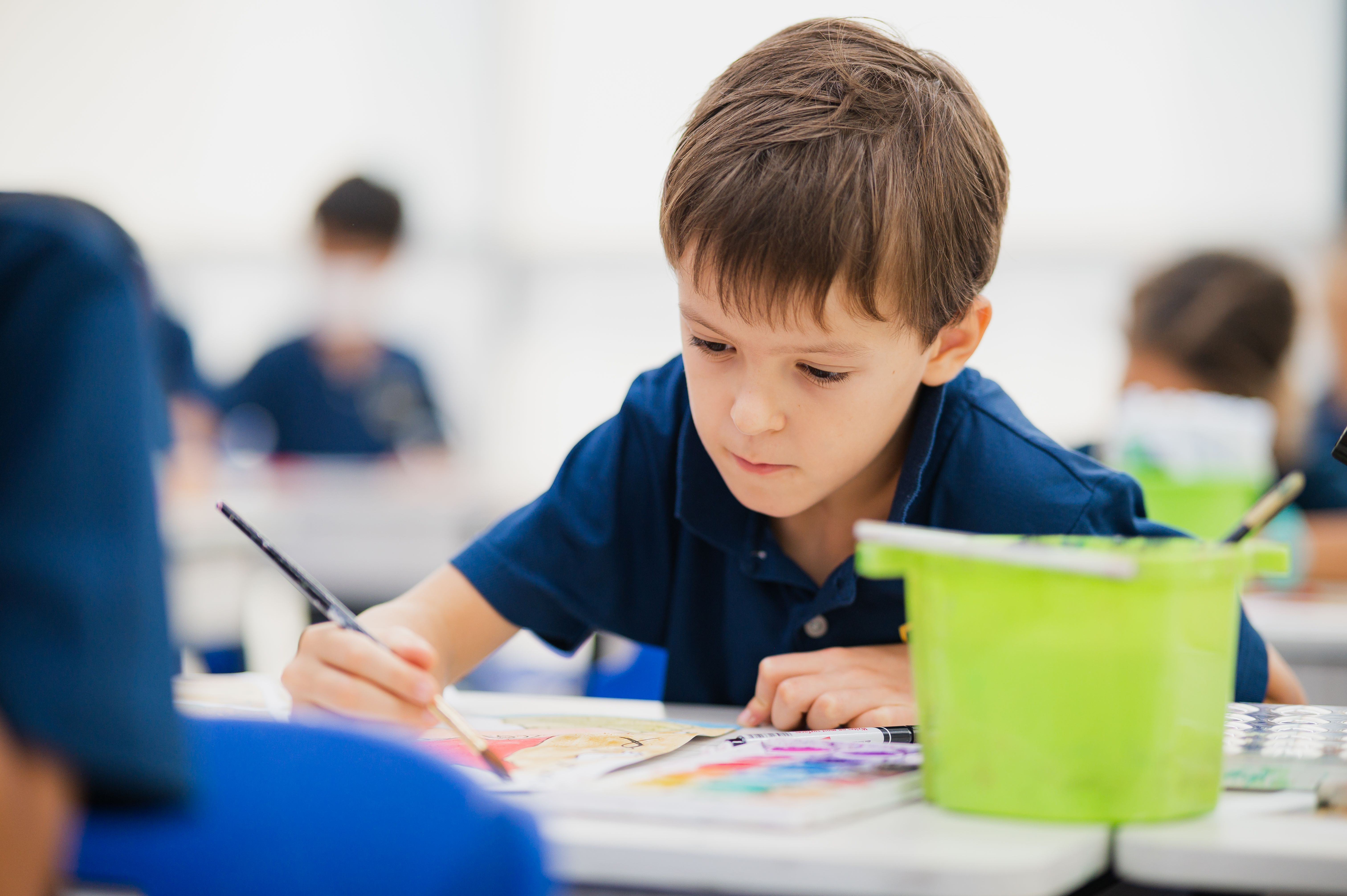 Young student at Invictus International School Singapore focused on a colourful painting activity during art class, seated at a desk with vibrant art supplies and a bright green container in the foreground.