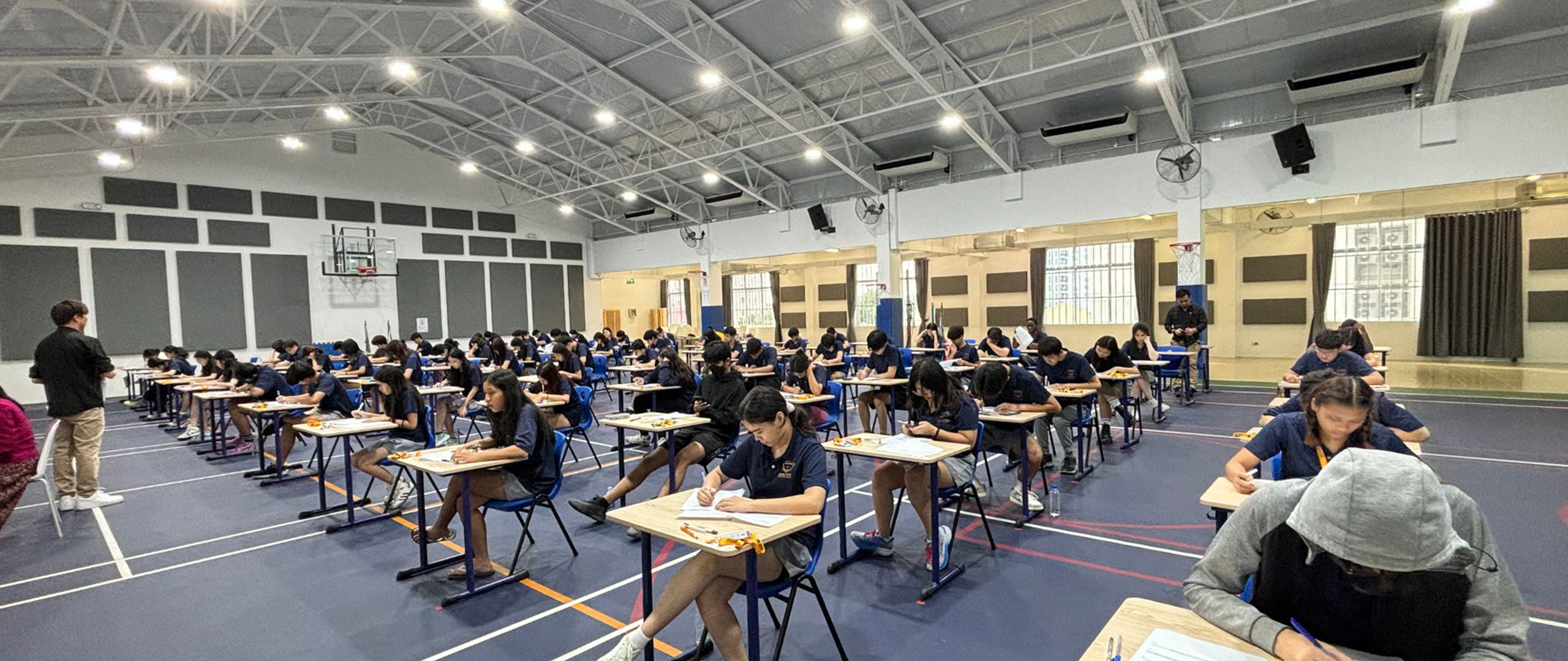 Secondary students at Invictus International School Cambodia seated in rows inside the campus multi-purpose hall, focused and writing during an official examination. The spacious, well-lit hall features high ceilings, modern lighting, and a calm, structured environment overseen by supervising teachers.