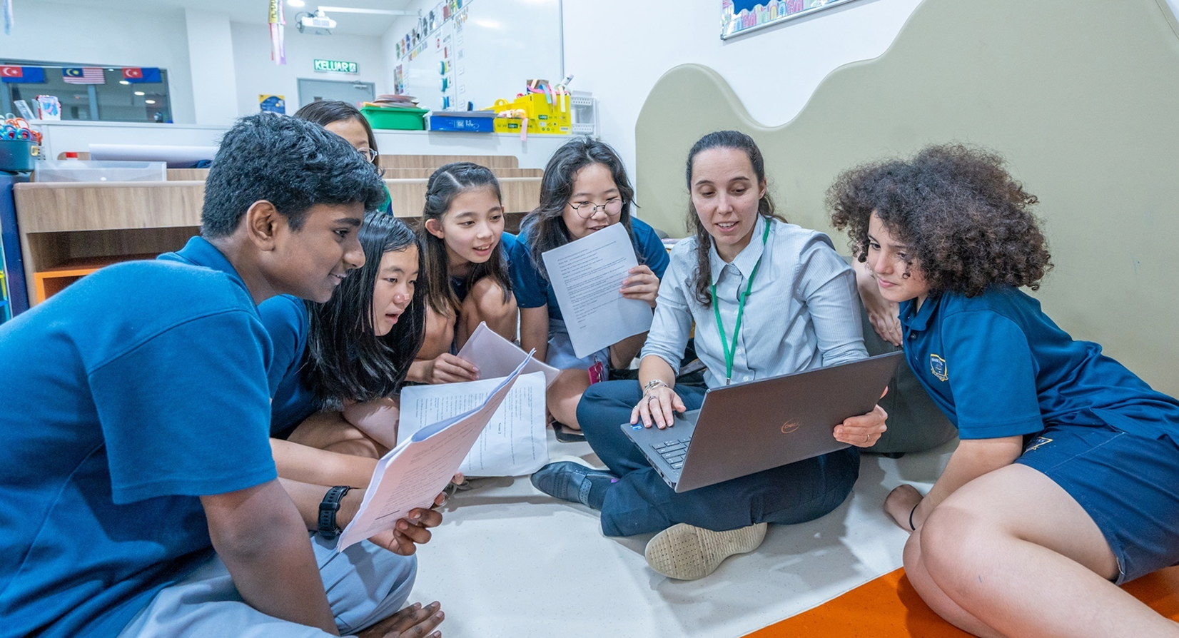 Teacher engaging a diverse group of Invictus Hong Kong students in collaborative learning on the classroom floor with worksheets and a laptop.