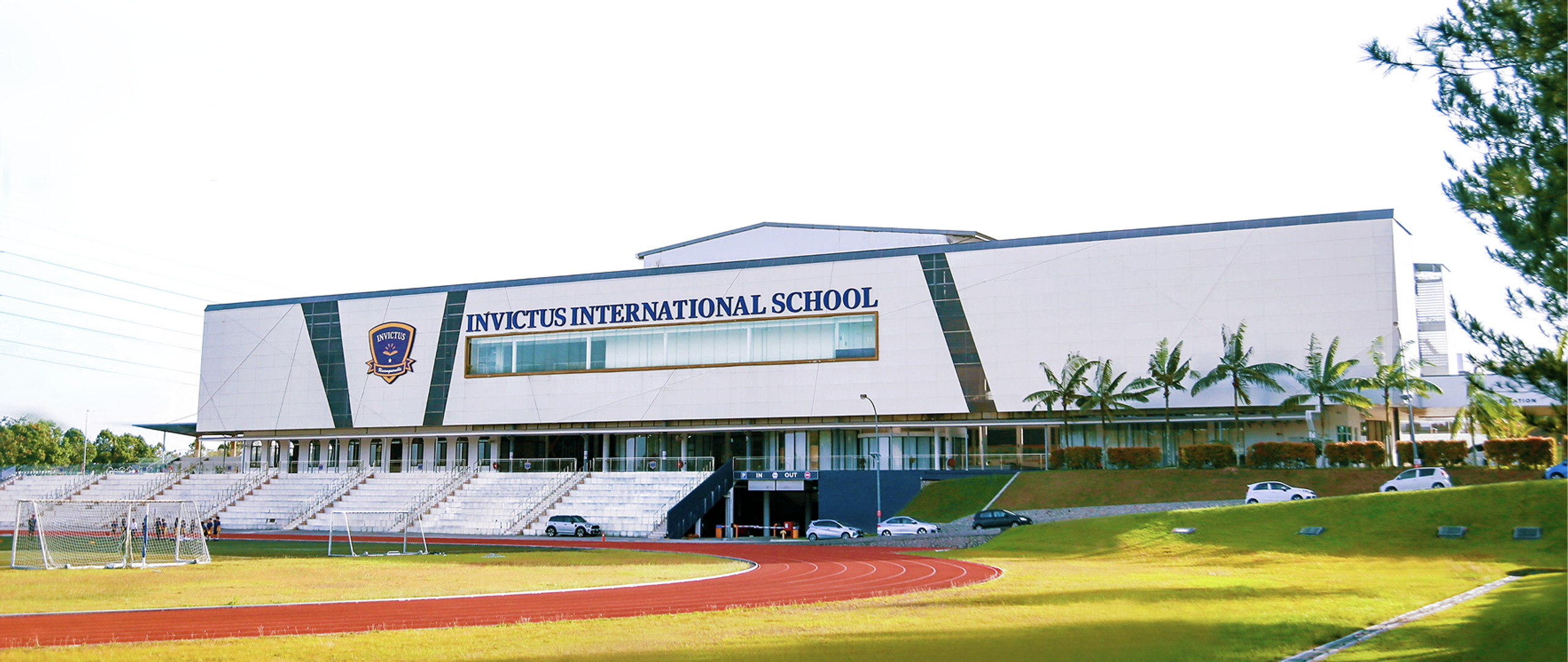 Aerial view of the Invictus International School Spring Hills campus, showing the modern main building façade, outdoor swimming pool, and adjacent sports track.