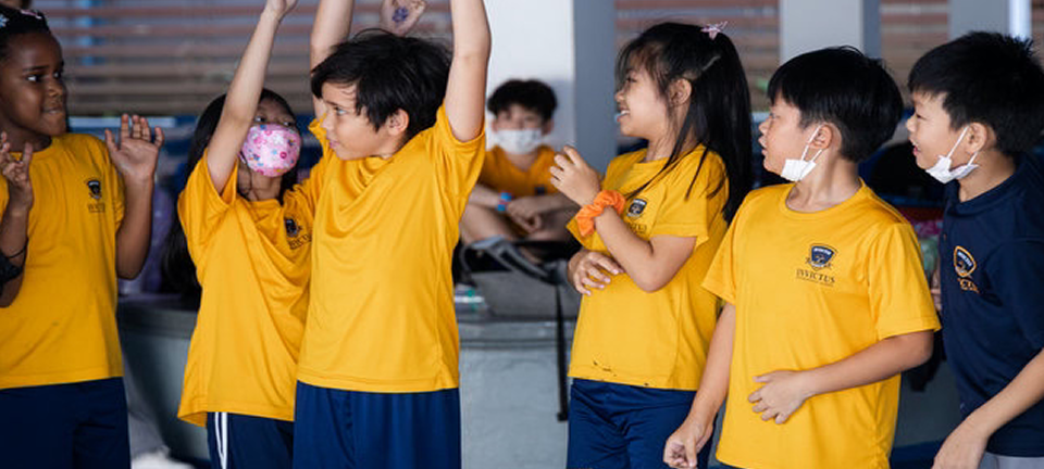 Primary students in yellow Invictus PE shirts standing in a row indoors, some with their hands raised in excitement while peers look on, all wearing school-branded uniforms.