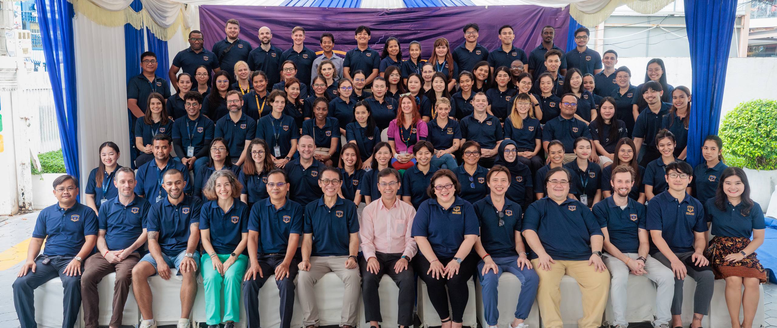 A group of Invictus International School Phnom Penh Year 4 students wearing masks and school uniforms enthusiastically perform in front of an art display and a teacher, with colourful values like "CARE" and "EXCELLENCE" painted on structural pillars in the background.