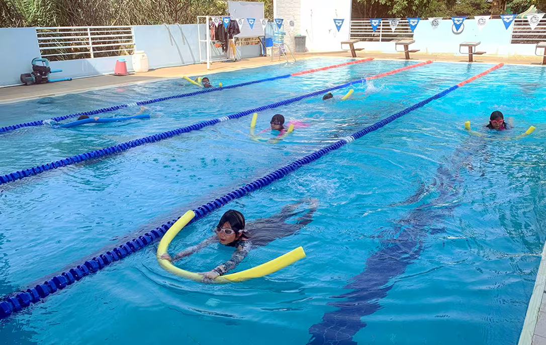 Children practicing swimming with float noodles in a clear blue outdoor pool with lane markers at Invictus Pathum Thani, supported by swimming instructors.