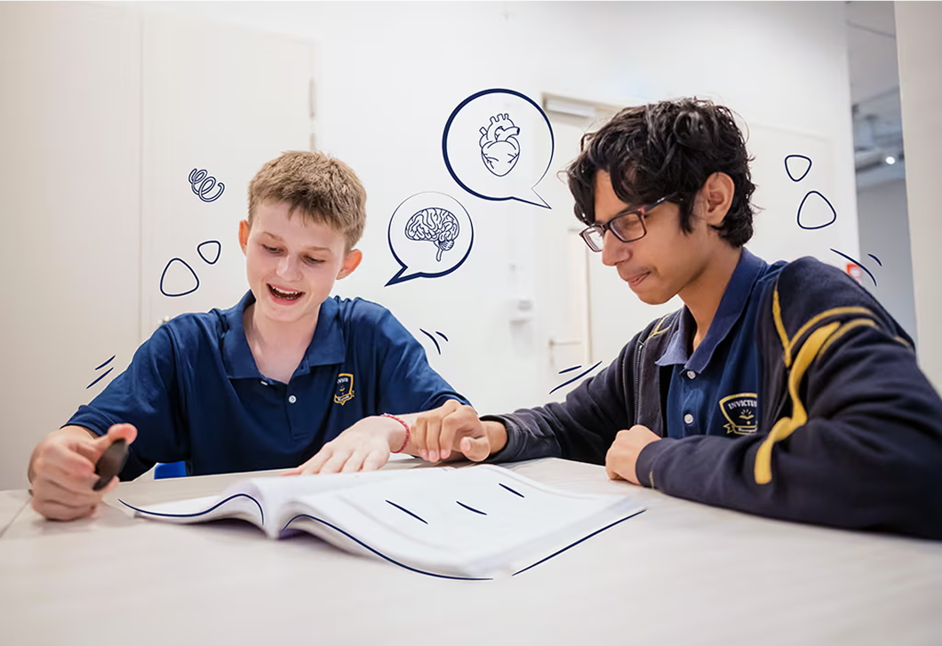 Two Invictus students in navy uniforms discussing a biology textbook, surrounded by illustrated thought bubbles showing the brain and heart.