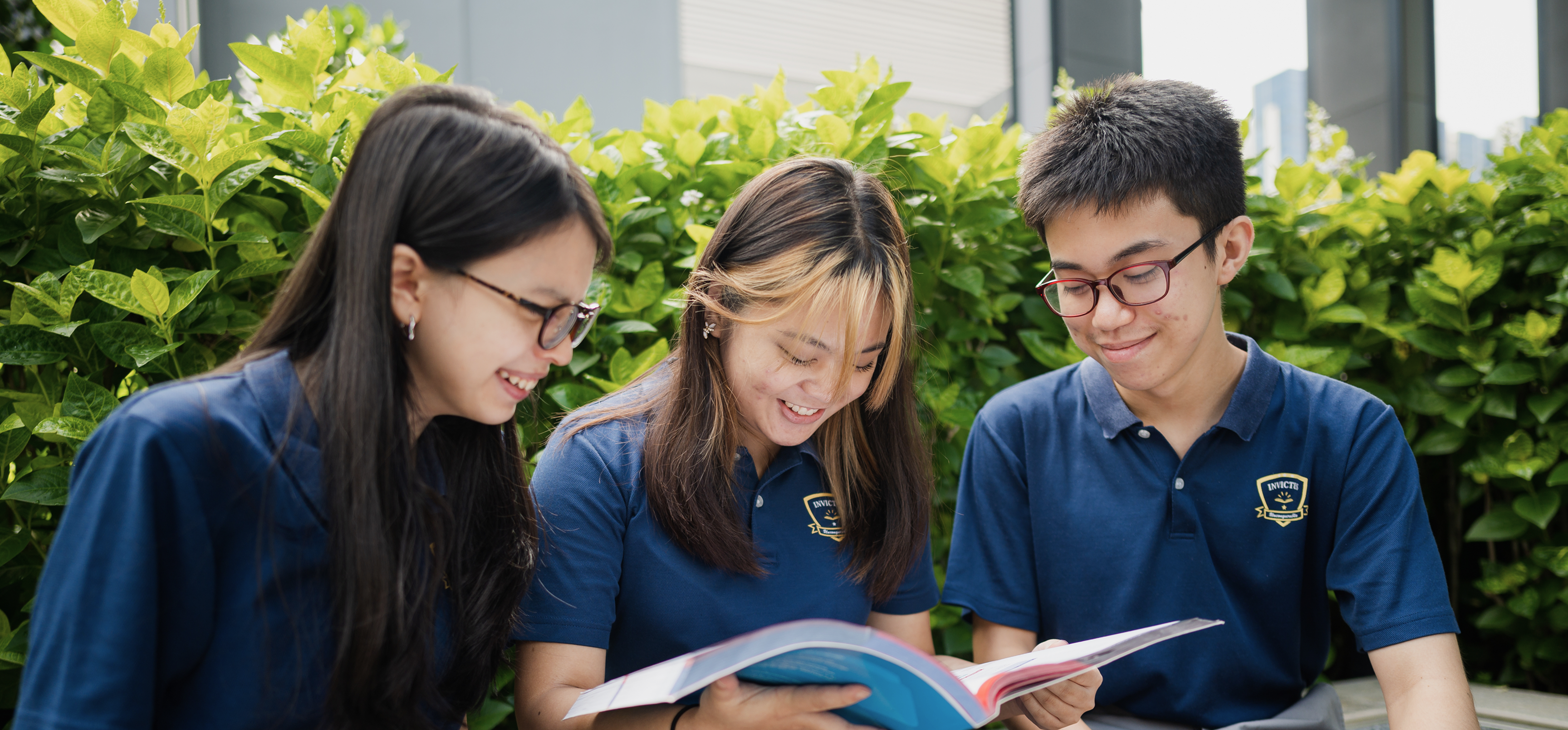 Three Invictus Secondary School students in uniform happily reading a book together outdoors on campus.