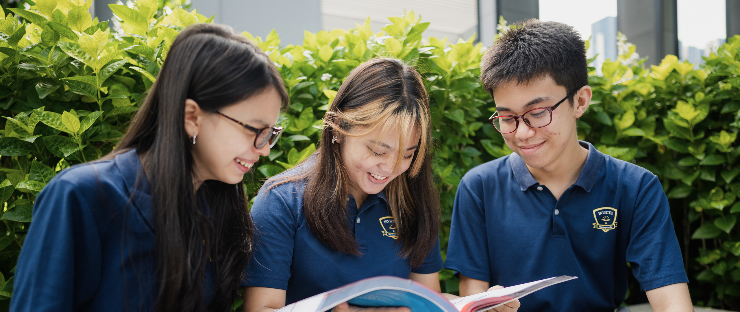 Three Invictus Secondary School students in uniform happily reading a book together outdoors on campus.