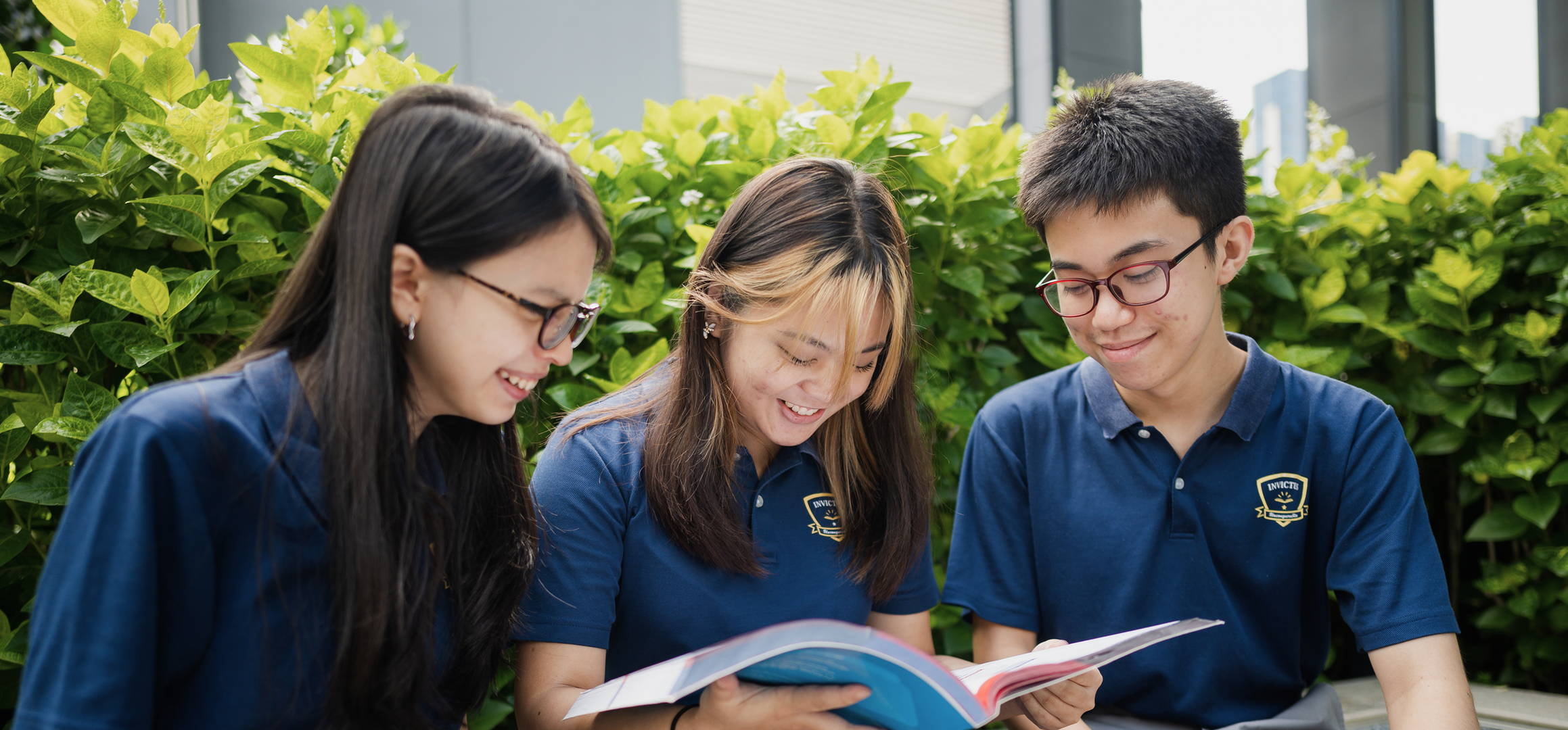 Three Invictus Secondary School students in uniform happily reading a book together outdoors on campus.