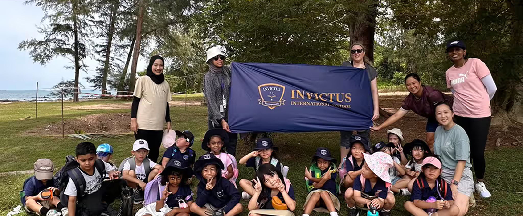 Students and teachers from Invictus Spring Hills on an outdoor learning trip by the sea, proudly holding the school banner in a scenic, natural setting.