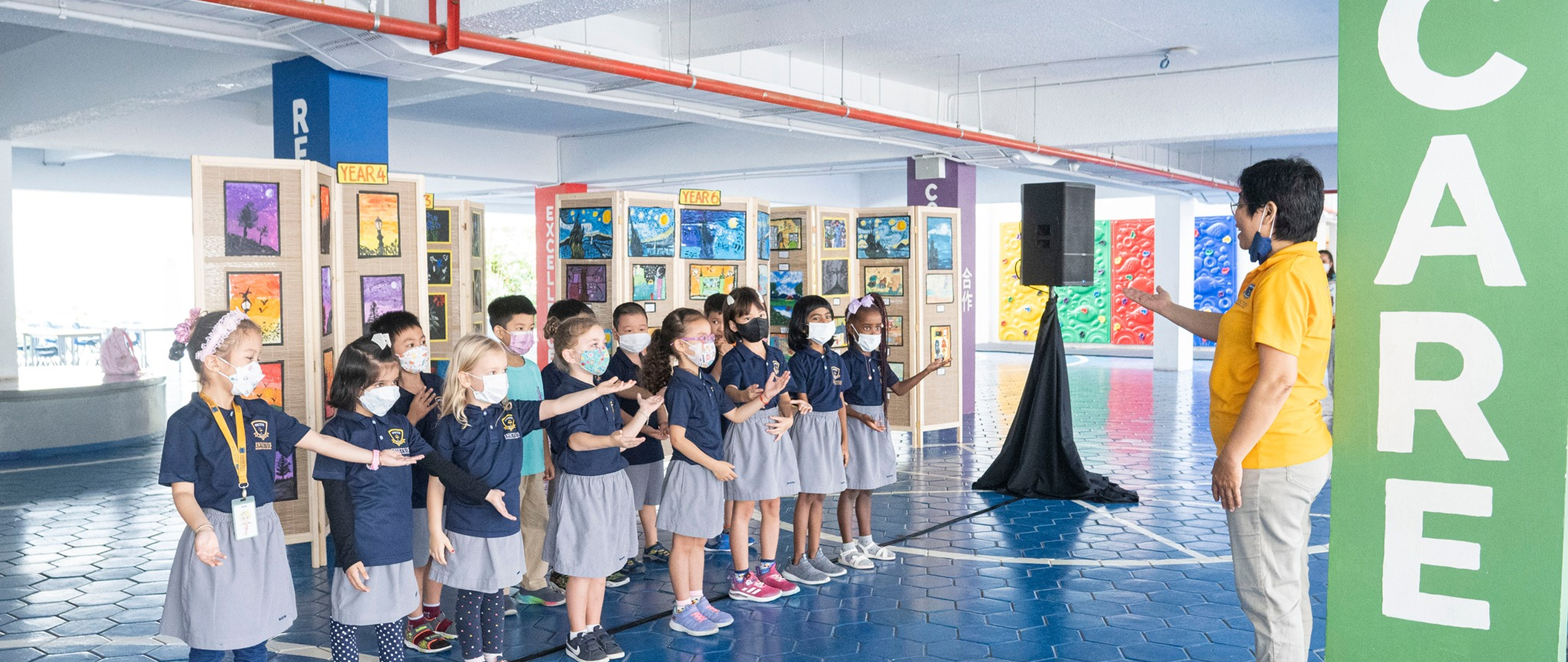 A group of Invictus International School Phnom Penh Year 4 students wearing masks and school uniforms enthusiastically perform in front of an art display and a teacher, with colourful values like "CARE" and "EXCELLENCE" painted on structural pillars in the background.