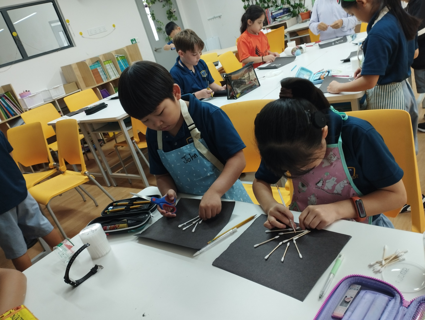 Two students in aprons at a classroom table arranging cotton-swab “rays” into a radial pattern on black paper, with art supplies scattered around.