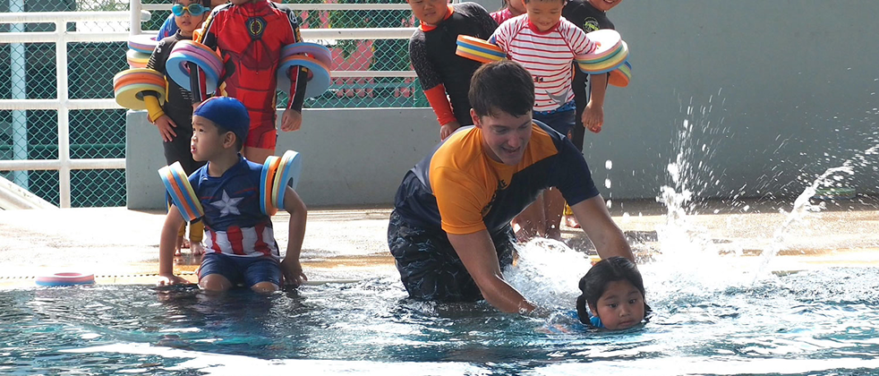 A teacher assists a young child during a swimming lesson at the Invictus Pathum Thani pool, while other children with floatation devices wait at the poolside.