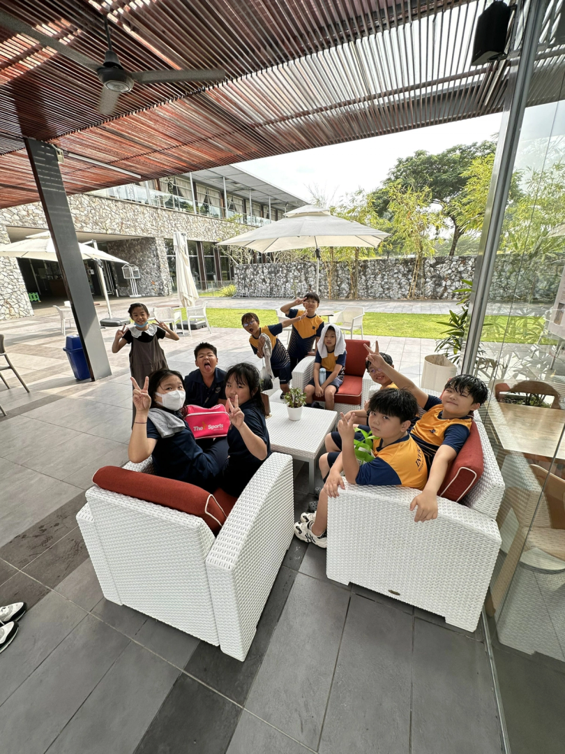 A group of primary students in school uniforms sit and stand around white wicker outdoor chairs and a small table under a covered patio, many flashing peace signs and playful expressions.