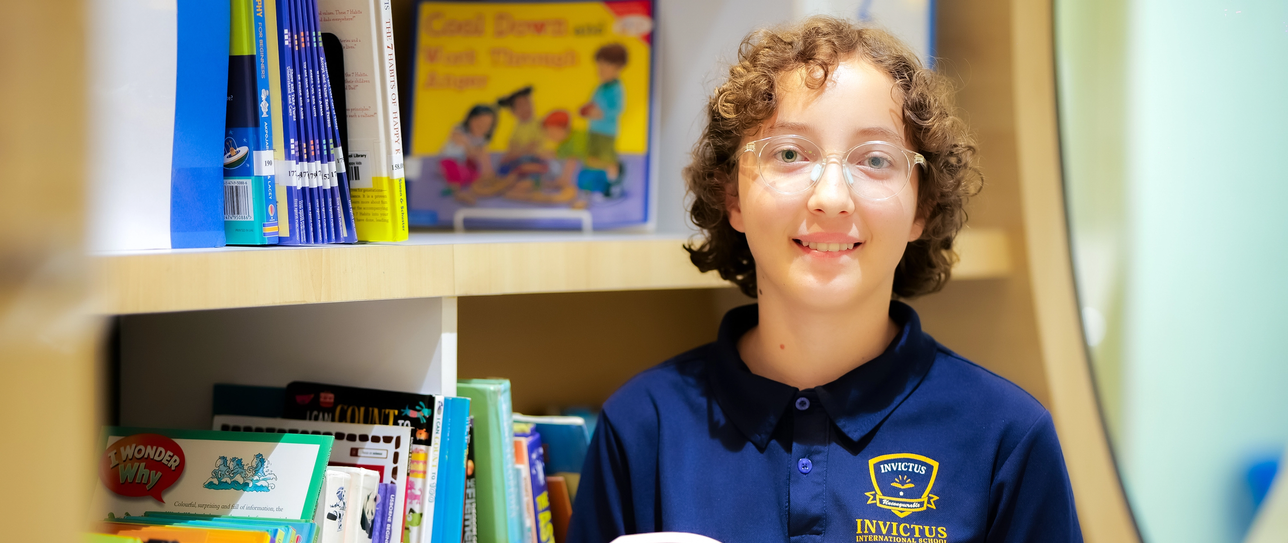A student smiles while holding a book in the Invictus school library, surrounded by colourful shelves, promoting a love for independent reading.