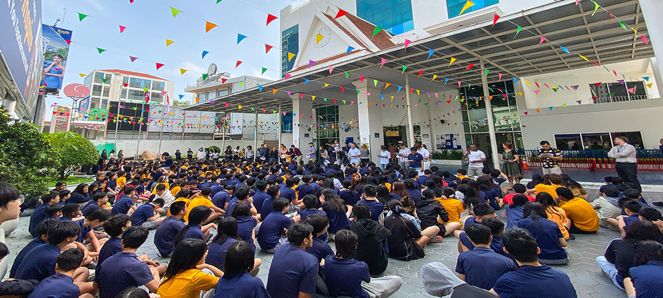 A large group of students in navy and gold uniforms seated on the courtyard paving under colourful bunting flags, listening to teachers speaking in front of the Phnom Penh campus entrance.