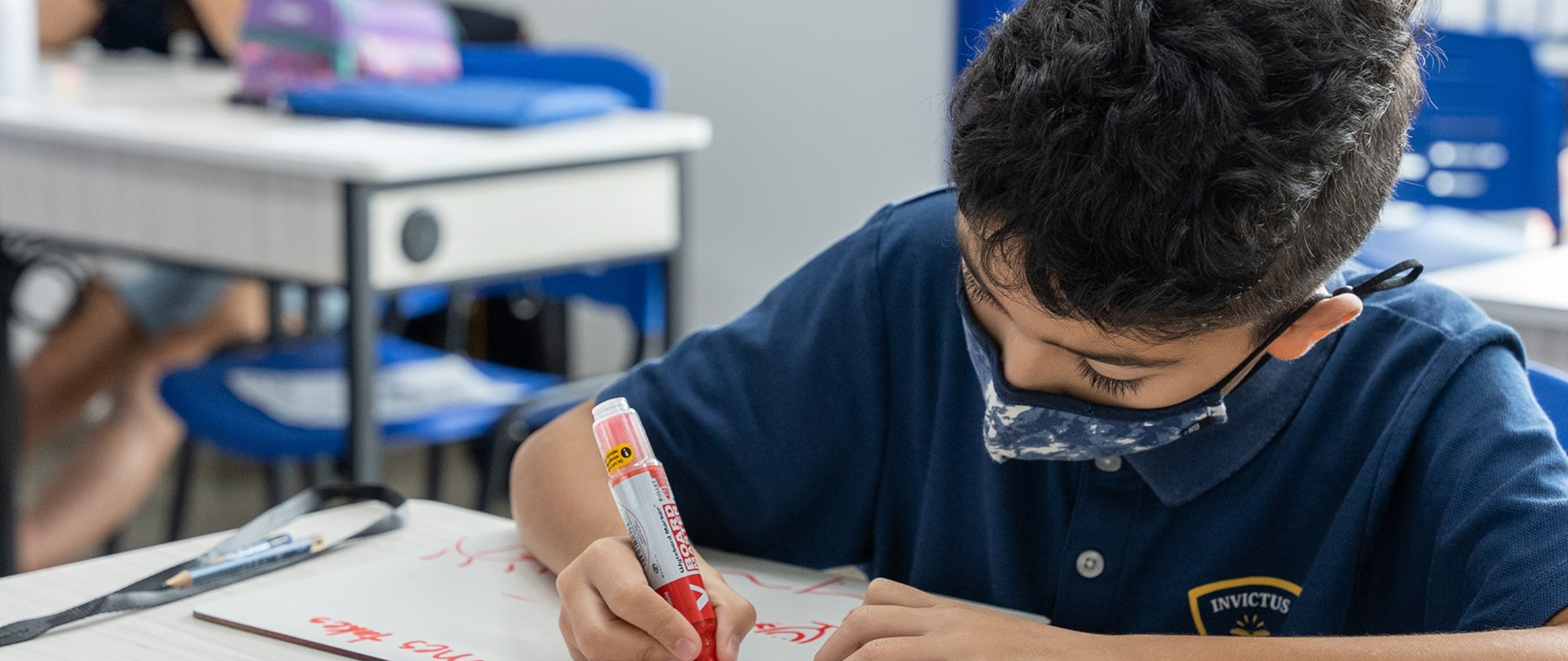A primary student at Invictus International School Thailand focuses on writing with a red marker on a whiteboard during class, wearing a face mask and school uniform.