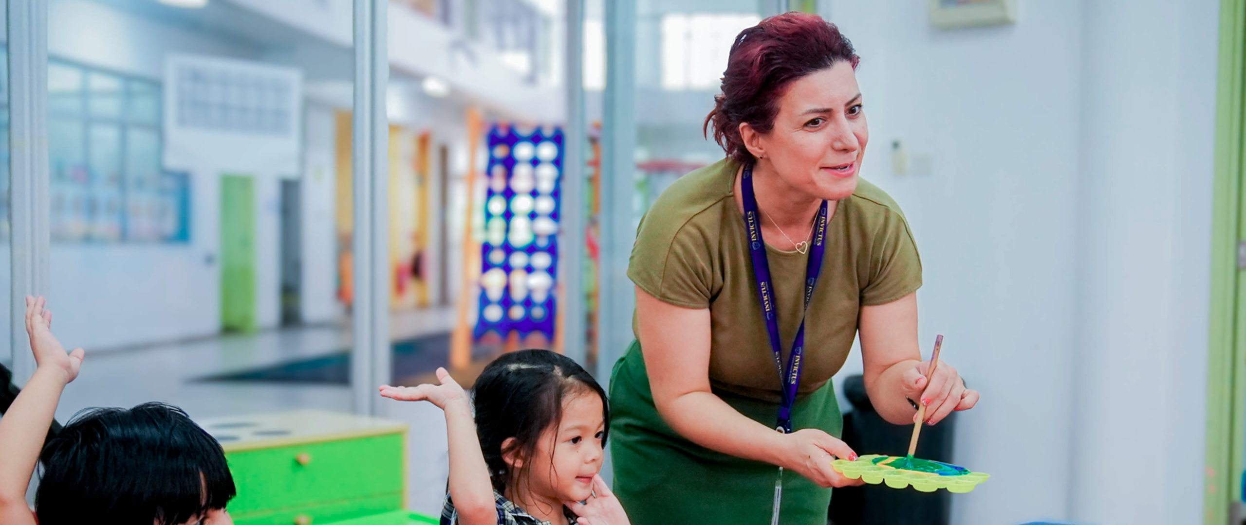 Teacher guiding young students with paint palettes in a bright Invictus classroom as they raise their hands during an art activity.