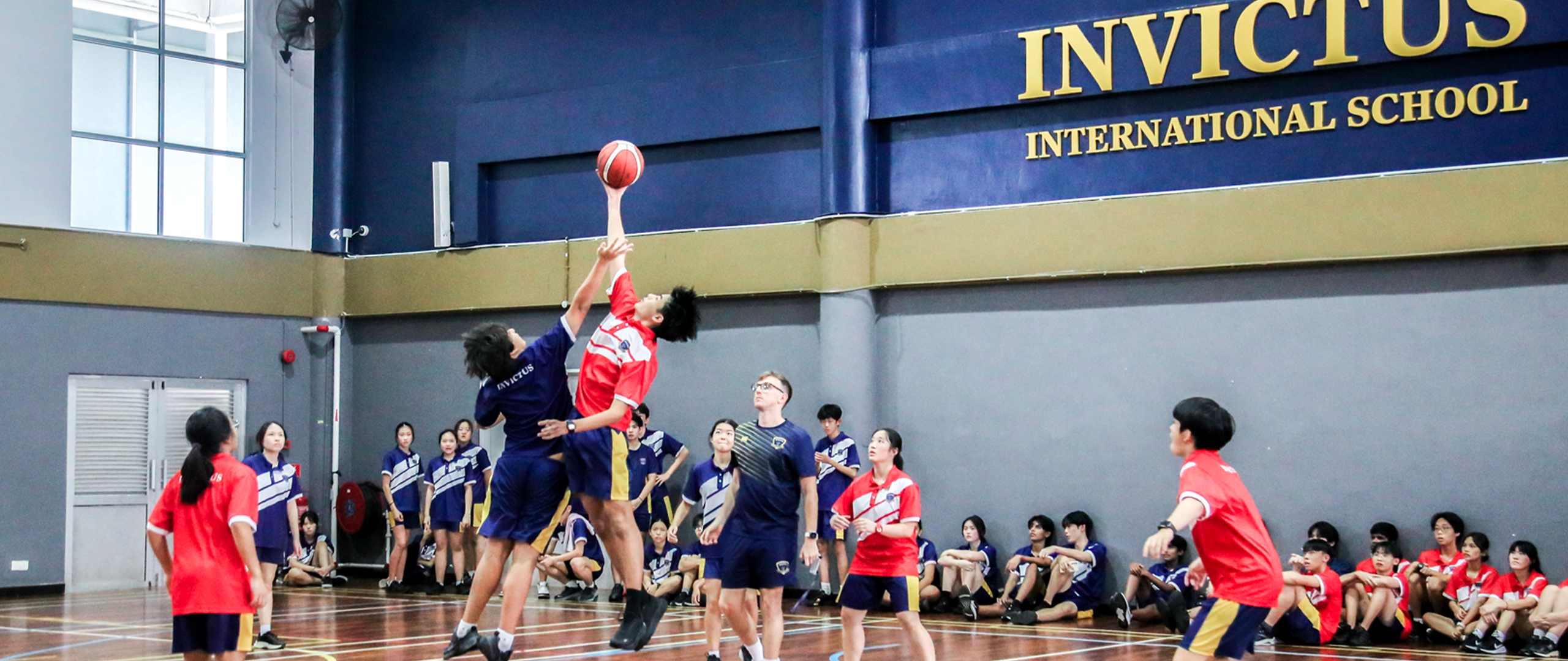 Two students leap for the basketball tip-off in the Invictus International School indoor gym, surrounded by teammates and spectators in school uniforms.