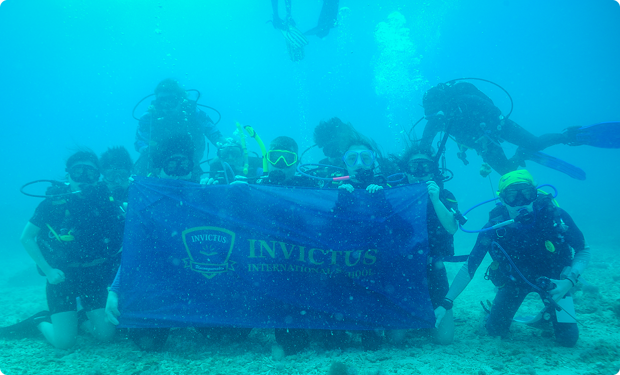 Invictus students scuba diving underwater, gathered around the school banner, participating in a marine learning activity.