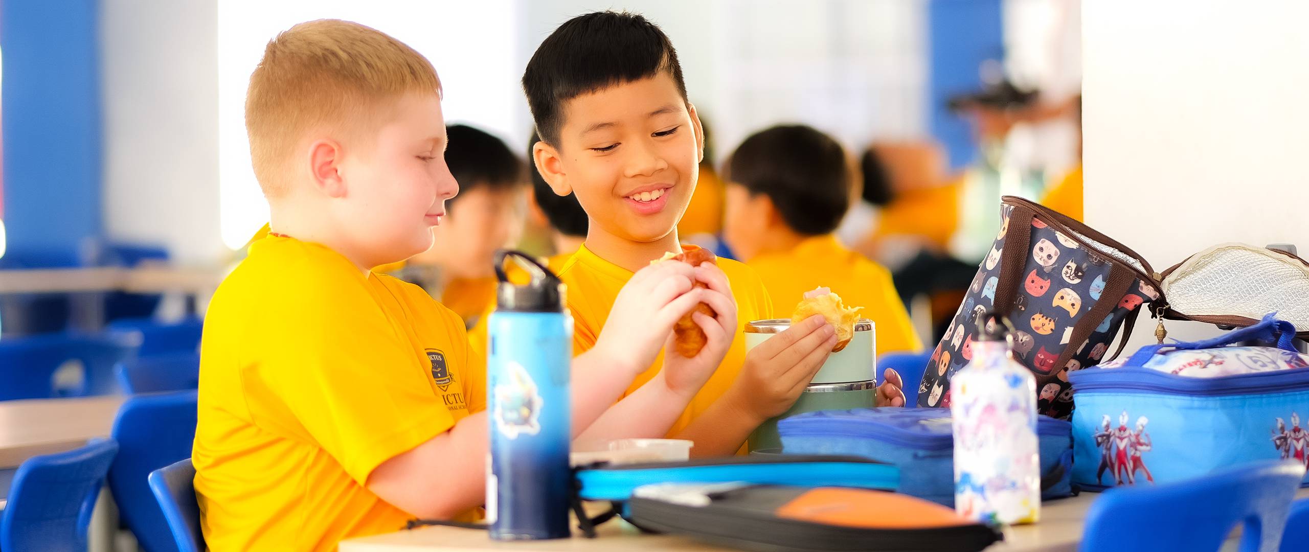 A group of Invictus International School Phnom Penh Year 4 students wearing masks and school uniforms enthusiastically perform in front of an art display and a teacher, with colourful values like "CARE" and "EXCELLENCE" painted on structural pillars in the background.