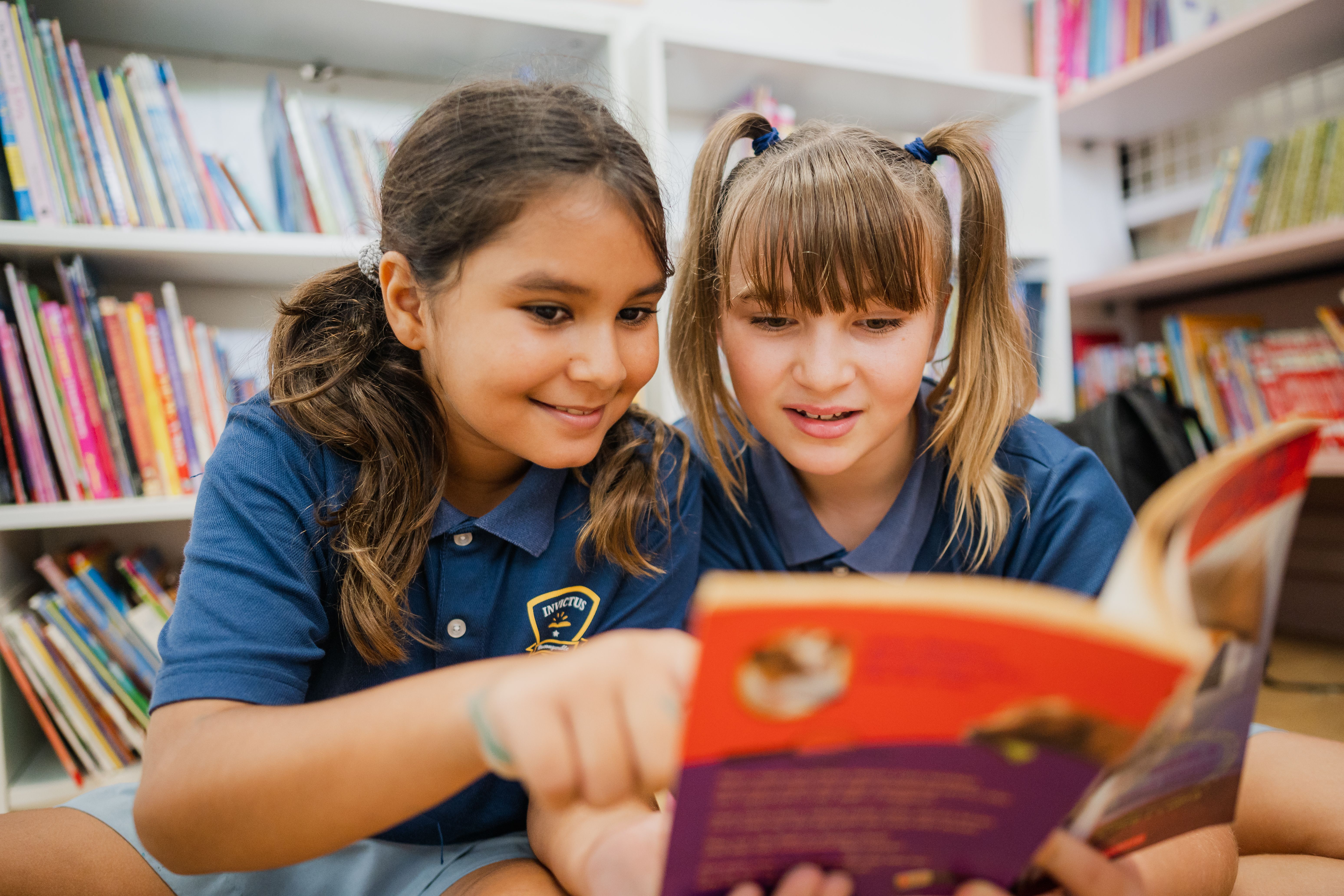Two young girls at Invictus International School reading a book together in the school library, smiling and engaged, surrounded by colourful bookshelves and dressed in Invictus uniforms.