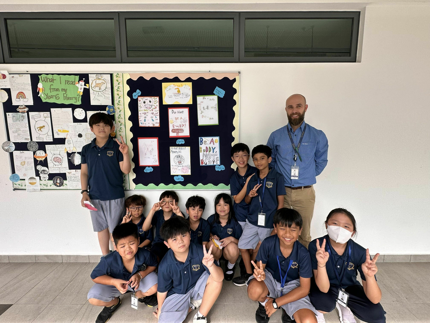 A male teacher stands with a group of uniformed primary students posing with peace signs in front of a wall display of their artwork.