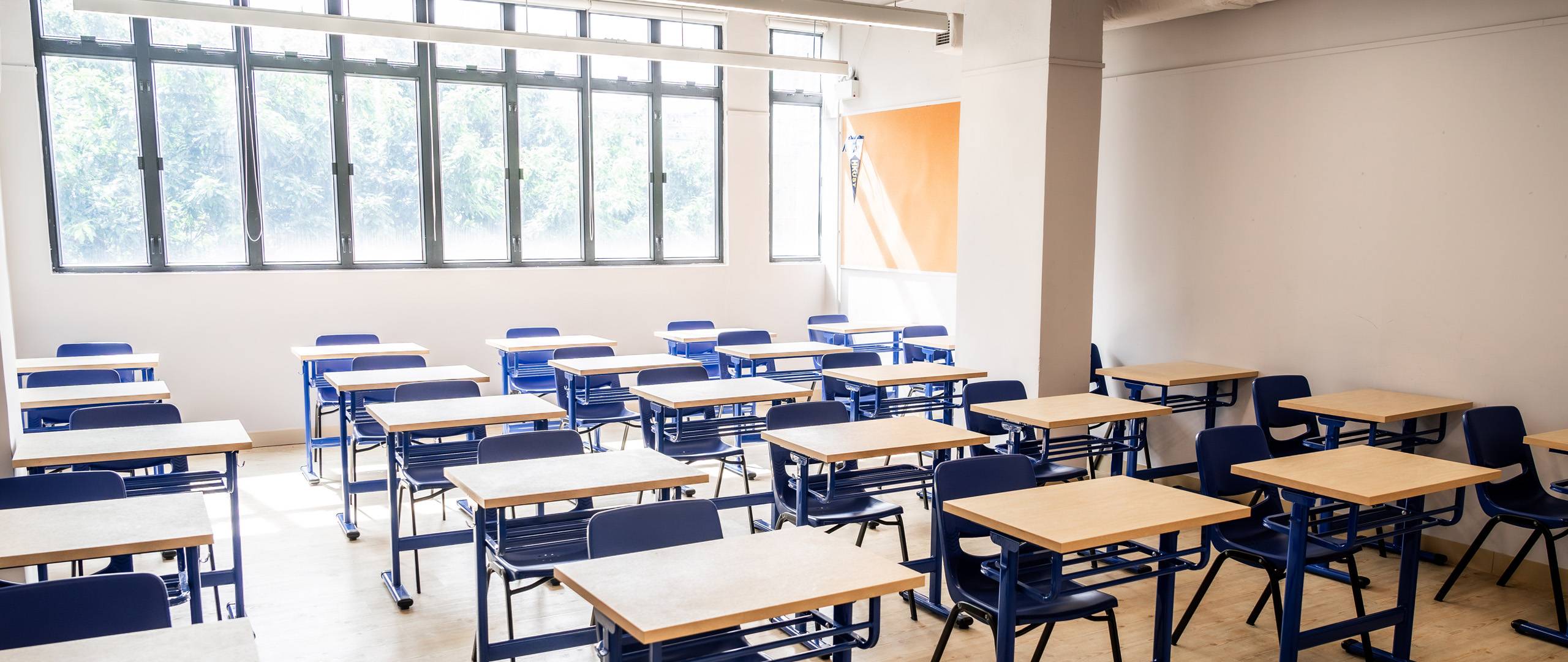Bright, sunlit classroom at Invictus School Hong Kong featuring neatly arranged blue chairs and wooden desks, with large windows and an orange bulletin board.