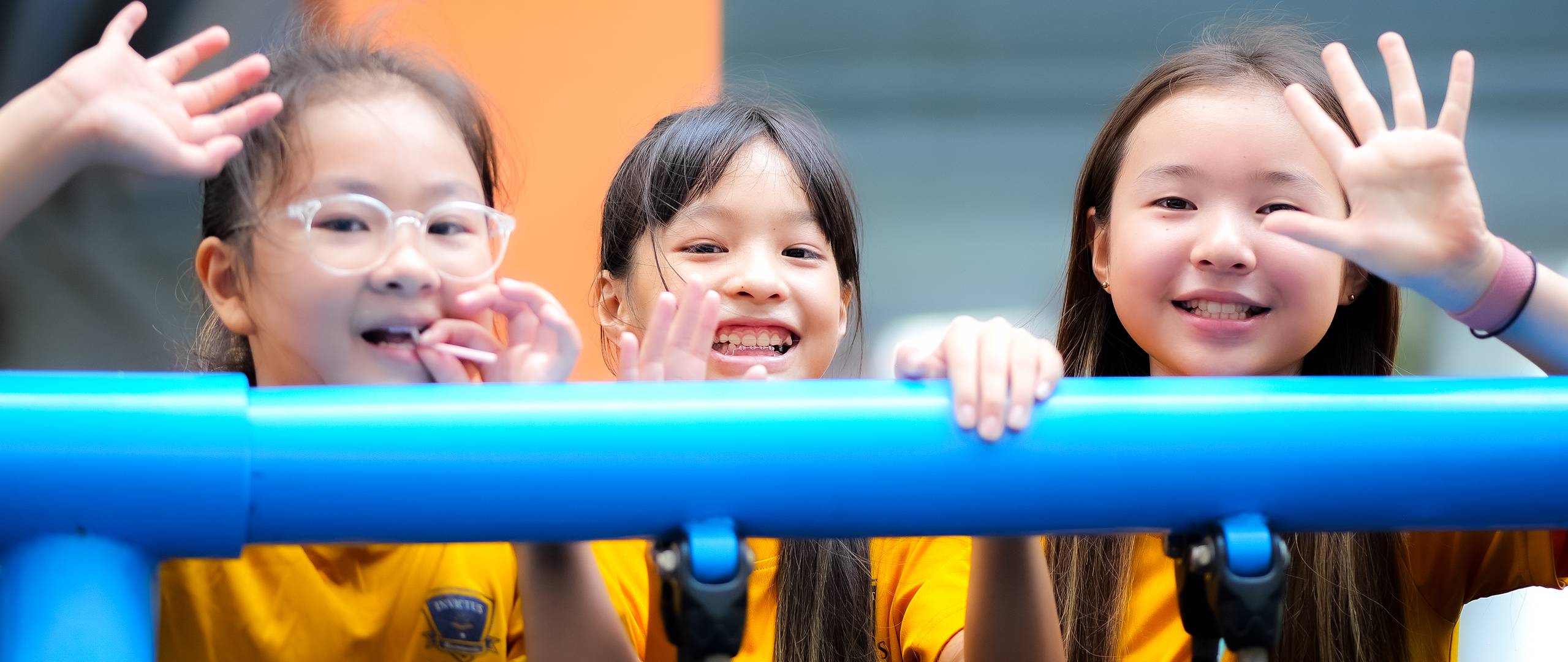 A group of Invictus International School Phnom Penh Year 4 students wearing masks and school uniforms enthusiastically perform in front of an art display and a teacher, with colourful values like "CARE" and "EXCELLENCE" painted on structural pillars in the background.