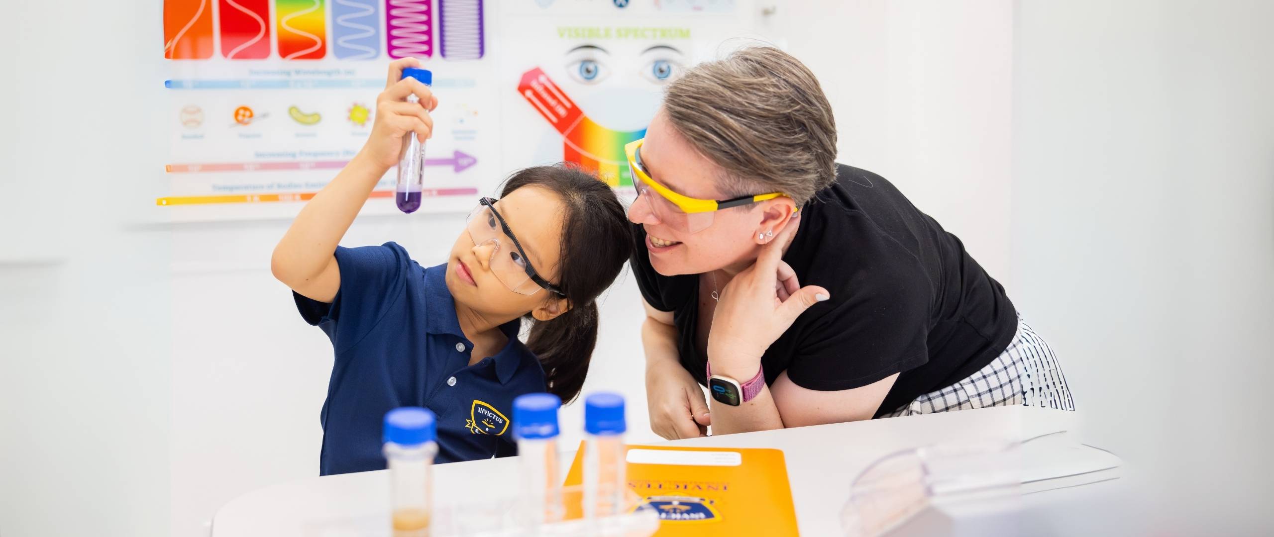 Invictus Bukit Timah student examining a test tube in a science experiment alongside a teacher wearing safety goggles in a bright classroom.