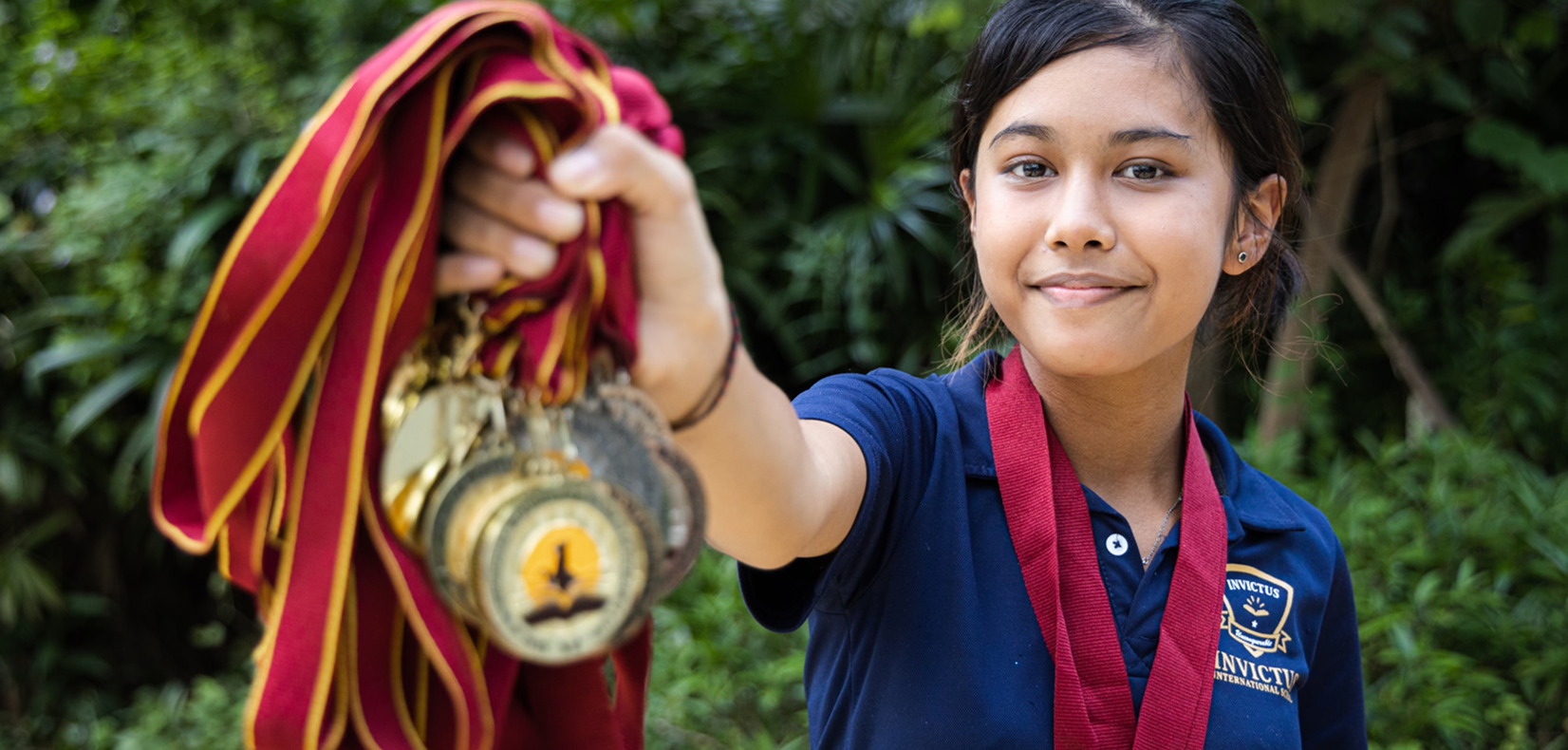 Invictus International School student proudly holding up multiple gold medals with a confident smile, showcasing academic or co-curricular achievement against a lush green background.