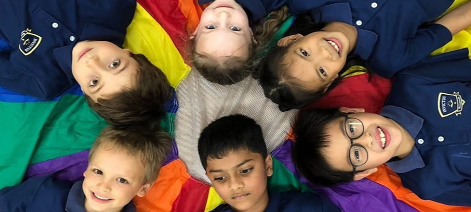 Six Early Years students in navy polos lying in a circle on a rainbow-coloured parachute, smiling and looking up at the camera.