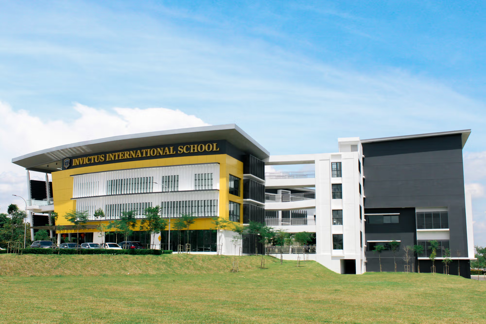 Wide-angle view of Invictus International School’s Horizon Hills Campus in Malaysia, featuring a modern building with bold yellow and black accents, large glass windows, and a spacious green lawn under a clear blue sky.