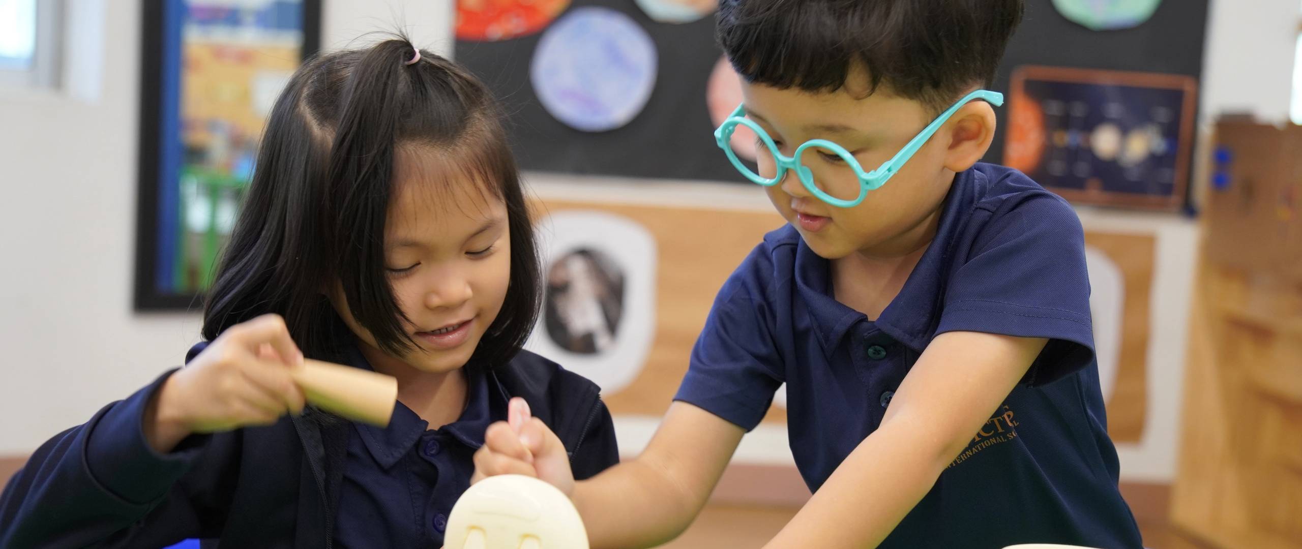 A group of Invictus International School Phnom Penh Year 4 students wearing masks and school uniforms enthusiastically perform in front of an art display and a teacher, with colourful values like "CARE" and "EXCELLENCE" painted on structural pillars in the background.