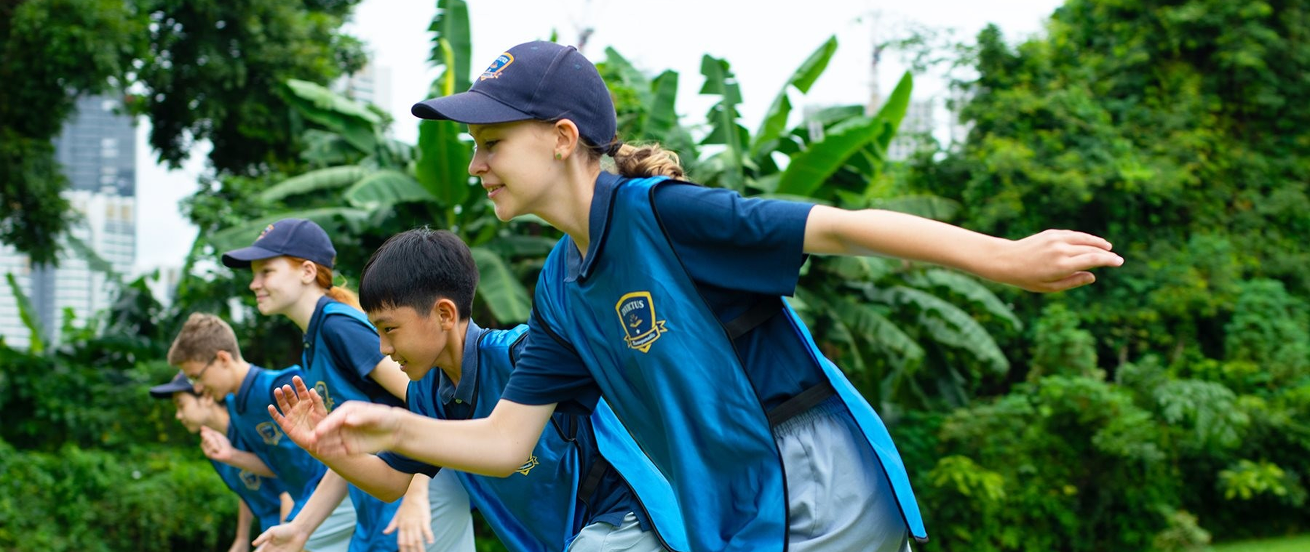 Invictus International School Thailand students participating in an outdoor sports activity, wearing blue sports vests and caps, surrounded by lush greenery and city skyline in the background.