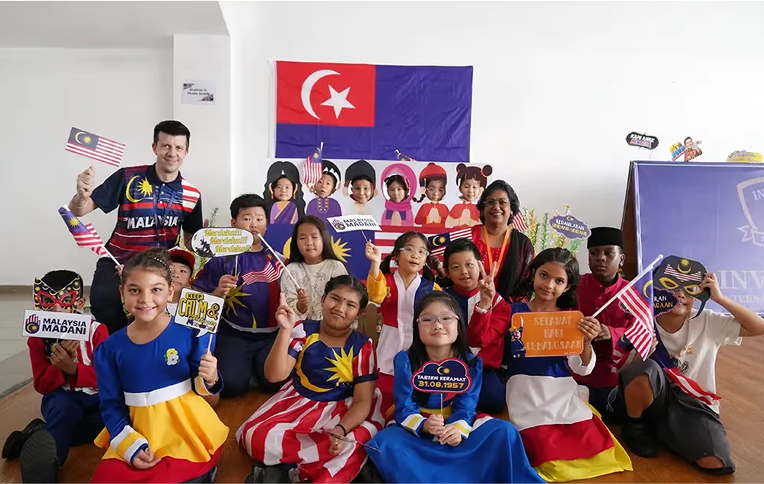 Students and teachers celebrating Malaysian culture during National Day with traditional attire, flags, and creative props in a festive classroom setting.