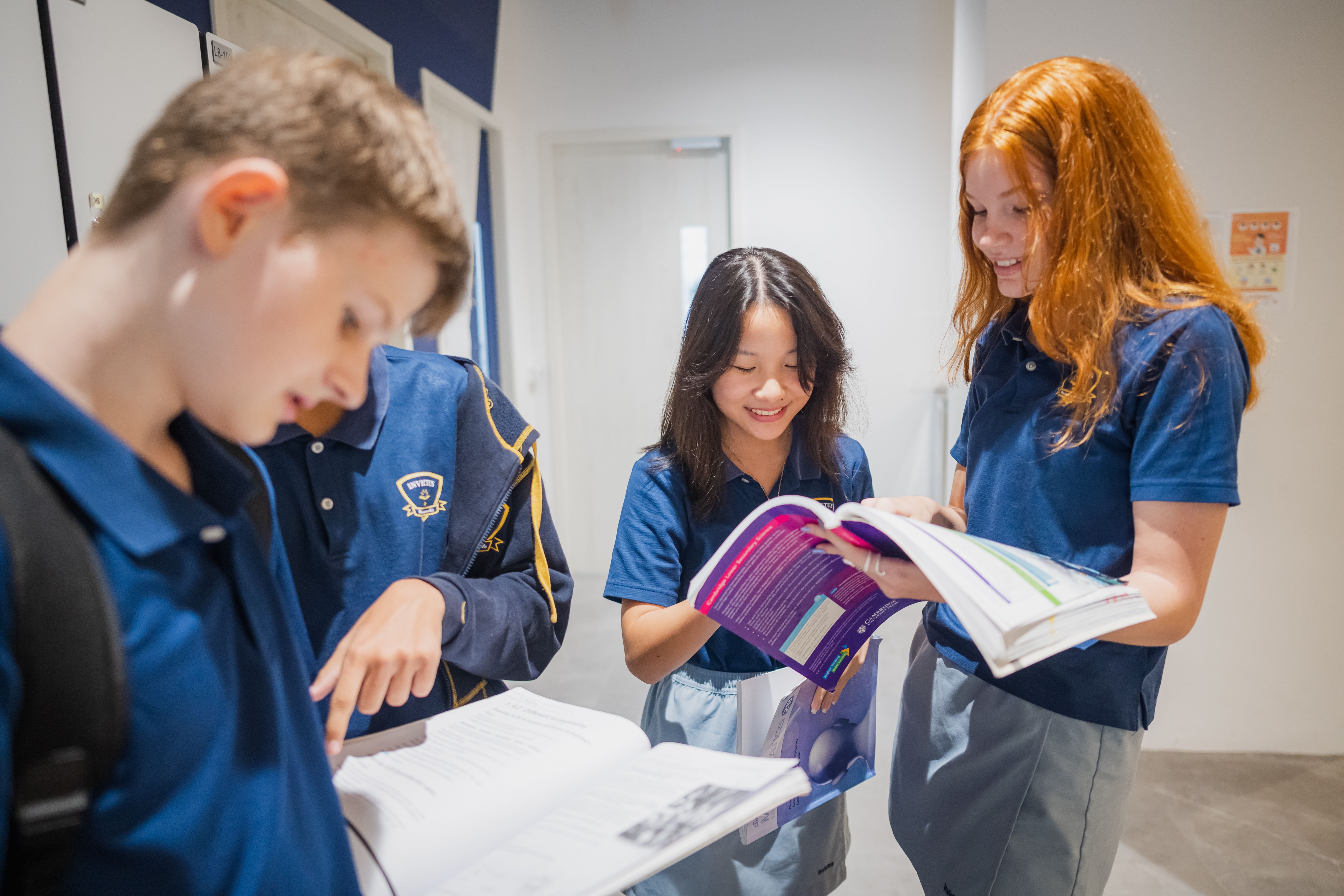 Two Invictus students in navy uniforms seated at a desk, one writing with a pen while the other follows along on a worksheet in a bright classroom.