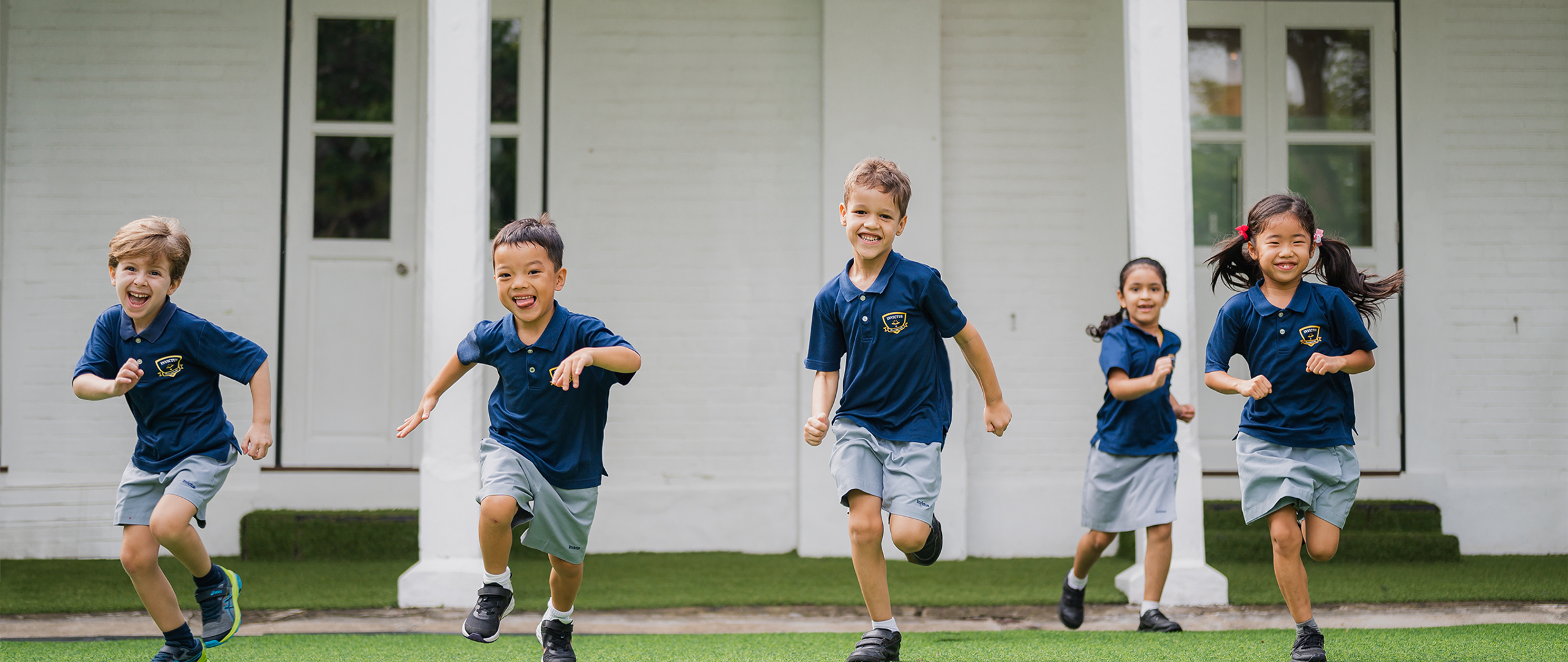 Five young students in Invictus International School uniforms joyfully running across a green field at the Dempsey Hill campus in Singapore, with a white colonial-style school building in the background.