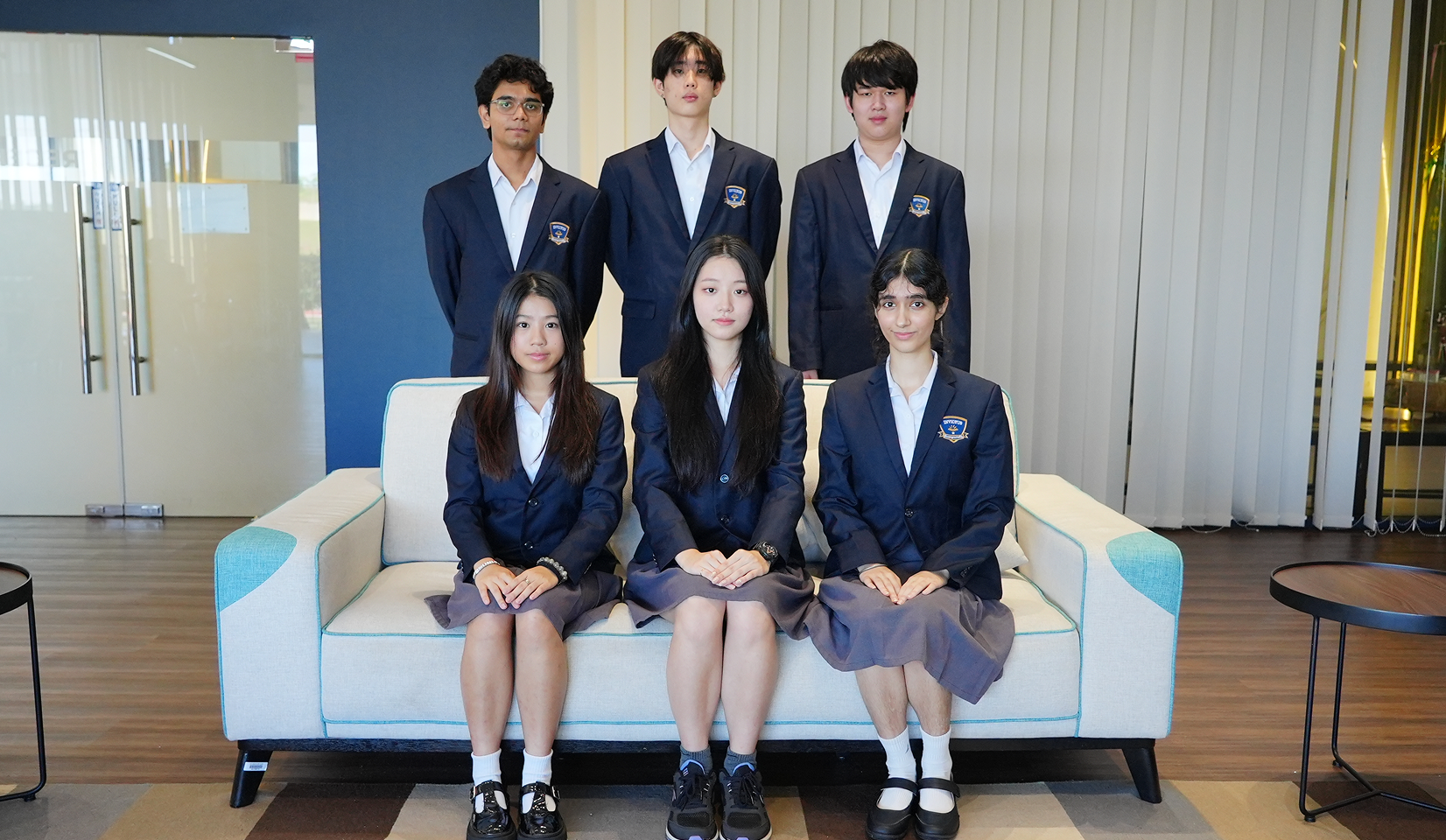 Six Invictus student scholars wearing navy blazers and white shirts: three standing behind and three seated on a light-coloured sofa, posing formally in a modern school lounge.