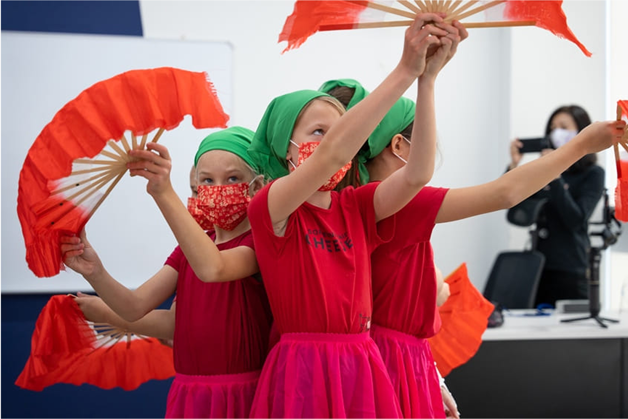 Young learners in bright red costumes and green headscarves performing a traditional Chinese fan dance in a school hall, with a teacher in the background recording their moves.